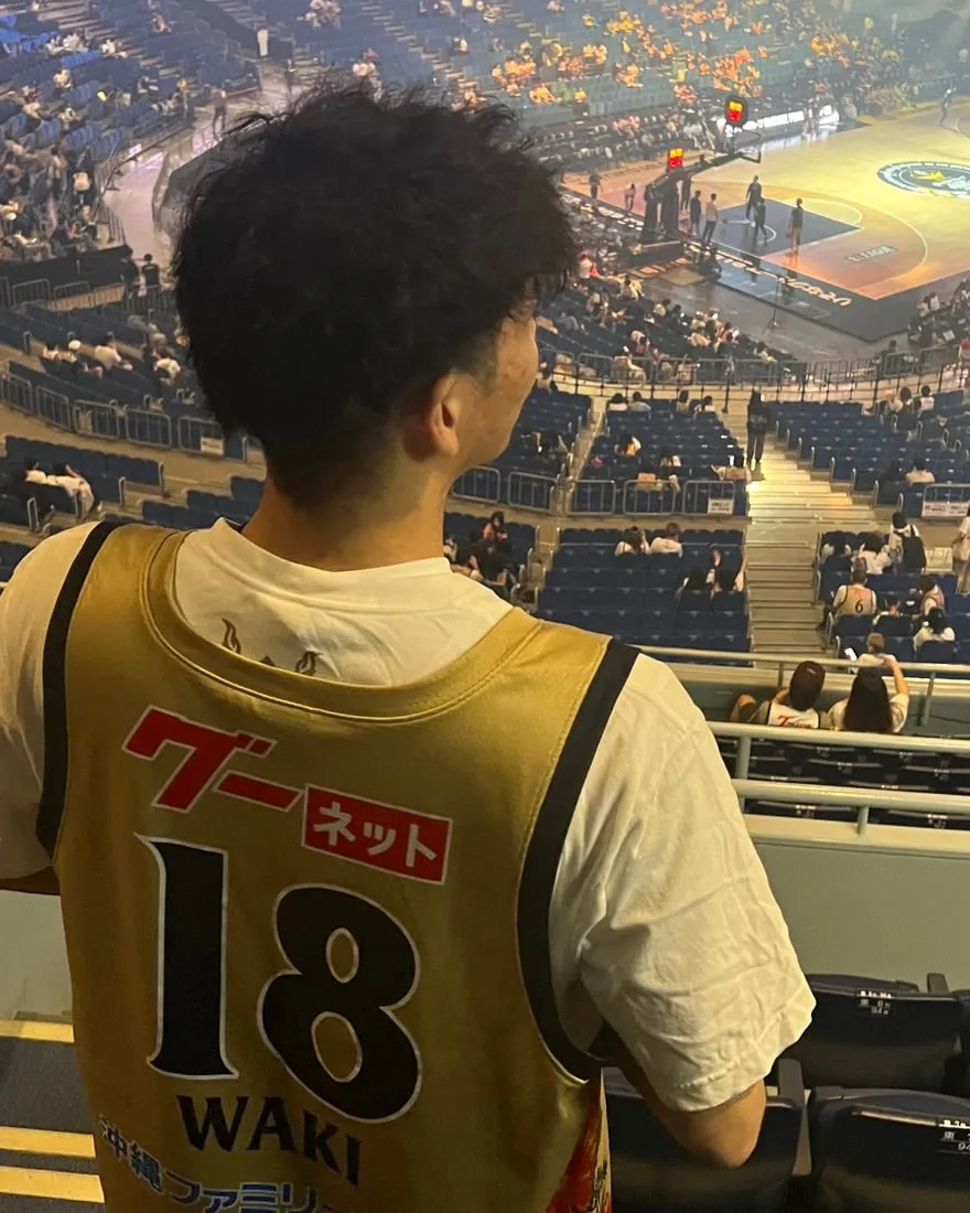 バスケットボールの試合会場の客席から、背番号18のユニフォームを着てコートを見下ろす後ろ姿